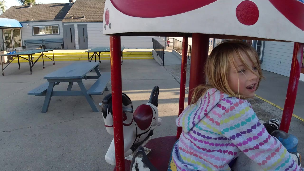 The child remains close to camera on the horse ride in Pismo Beach, with a gray picnic table, yellow curb, railing, and building doors visible in the background.
