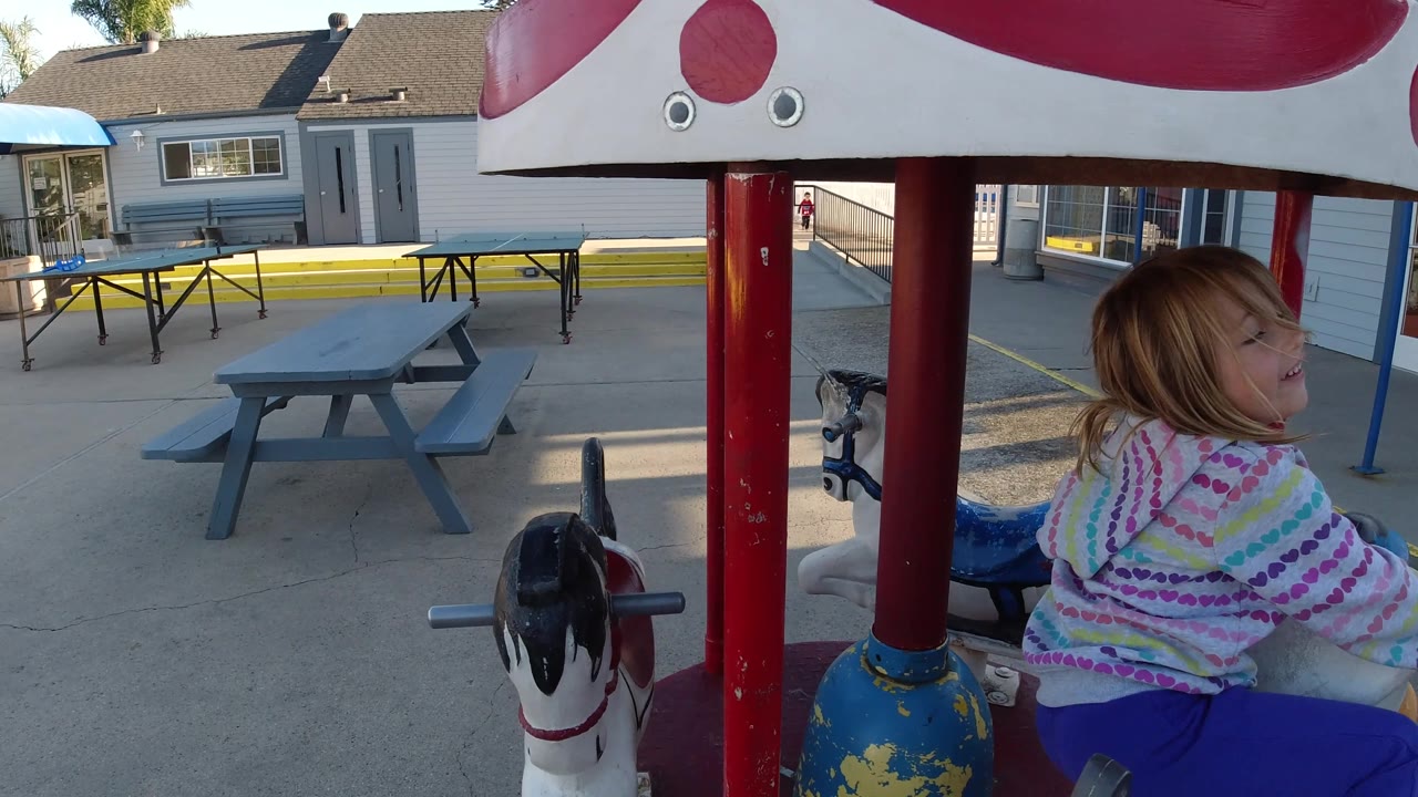 The child turns farther to the right on the carousel-style horse ride in Pismo Beach, framed by red poles with painted horses and a shaded paved courtyard behind.