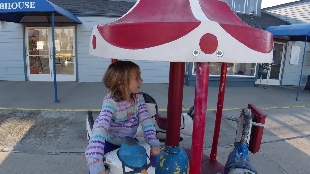 In Pismo Beach, California, the girl turns to the side on the carousel-style ride, with the red and white canopy above and building doors and windows behind her.