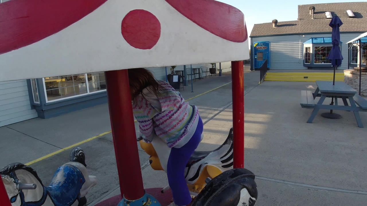 A young girl stands on a child-sized carousel under a red-and-white canopy in Pismo Beach, with painted ride animals, red support posts, storefront windows, and pavement behind her.