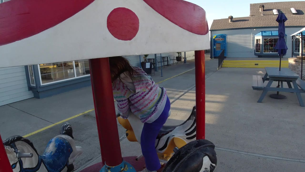 The girl leans forward on the small carousel in Pismo Beach, framed by red poles, chipped blue and black ride animals, a gray building, and an outdoor picnic table.
