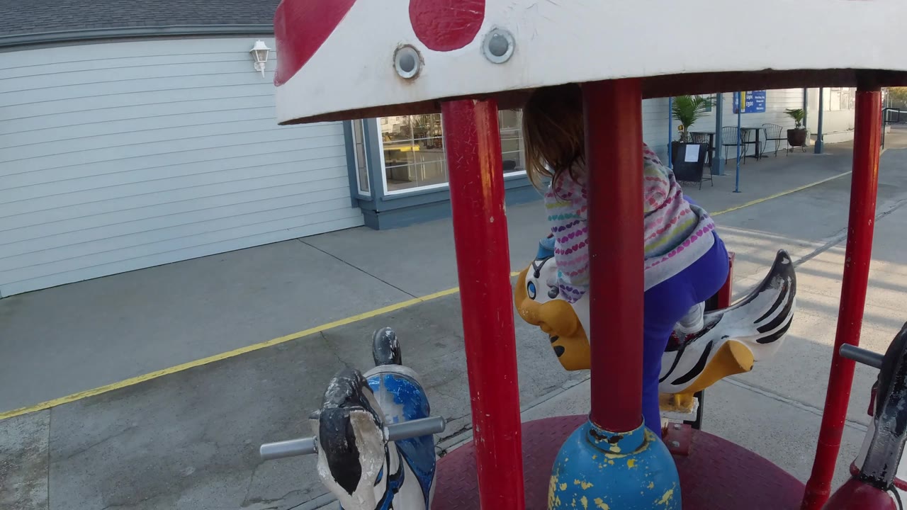 The girl appears near the right side of the carousel in Pismo Beach, with the canopy edge overhead, red poles in front, a blue ride figure, storefront siding, windows, and marked pavement behind.