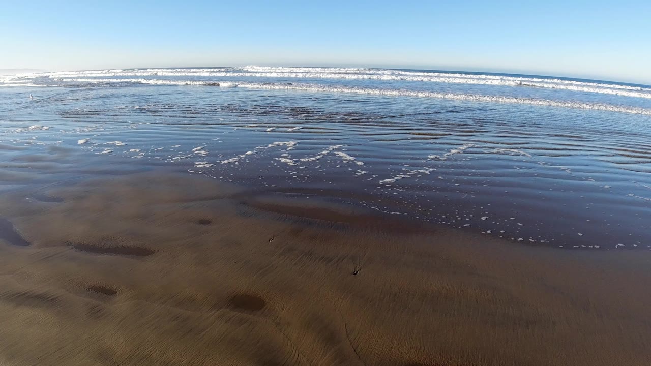 Low view of wet beach sand, shallow water, and white surf under blue sky.