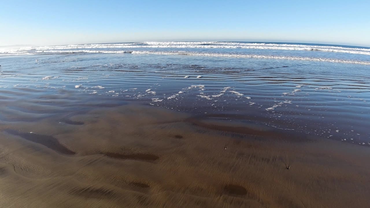 Pismo Beach shoreline shows brown wet sand patches beneath shallow water, small foam clusters near midframe, and bright surf across the background, with no visible people.