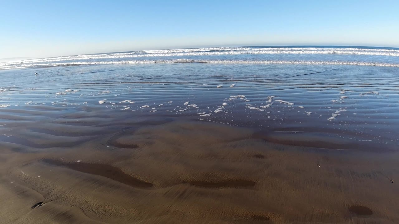 Pismo Beach surf sits farther back behind a wide sheet of shallow water and smooth wet sand, with scattered foam and no visible people.