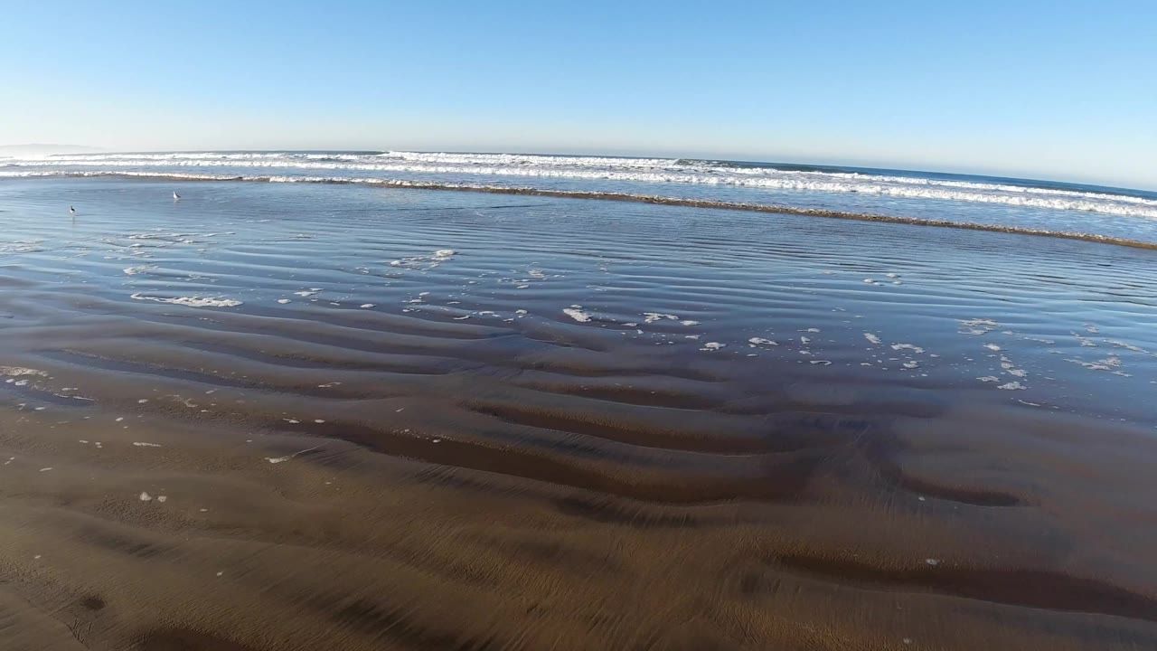 Pismo Beach shoreline appears as reflective wet sand with thin foam streaks in the shallows, breaking waves in the distance, and no visible people.