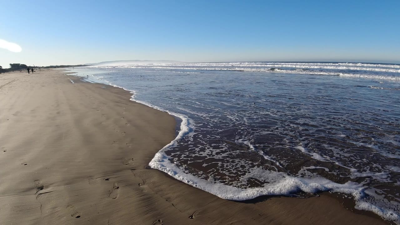 Wide Pismo Beach shoreline at low tide with wet sand in the foreground, a curved edge of white foam, low ocean waves, and small distant people near the left horizon.