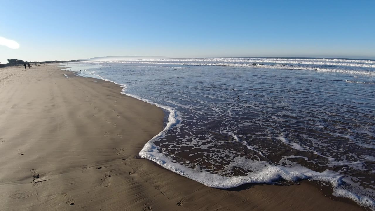 Curving surf foam on wet beach with distant figures near the left side.