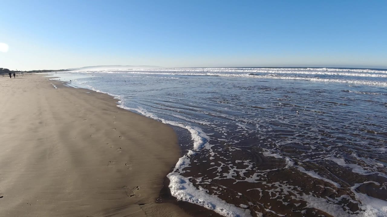 Wide Pismo Beach low-tide view with reflective wet sand across the right foreground, white wave bands offshore, and two small people walking or running near the left beach edge.