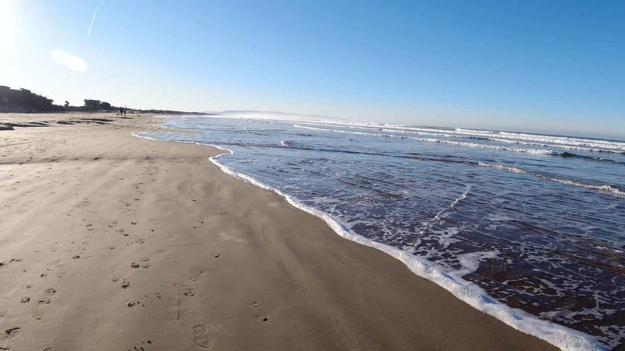 Pismo Beach shoreline under a clear blue sky, with wet sand in the foreground, white surf foam curving in from the right, and tiny distant people near the left background.