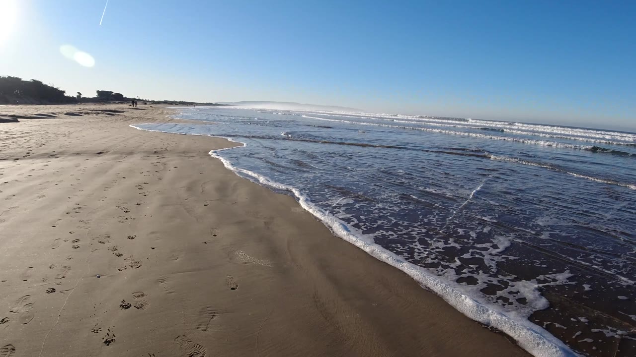 Pismo Beach shoreline with a broader sheet of shallow water in the foreground, white foam tracing the edge of the tide, and the beach stretching toward tiny people in the distance.