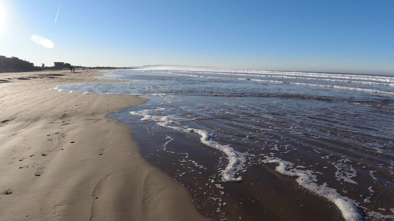 Pismo Beach foam trails spread across dark wet sand as low ocean waves roll in the distance, with no visible people in the frame.