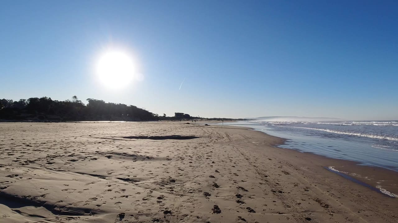 Pismo Beach shoreline under a high bright sun, with textured sand in front, waves rolling in from the right, and no visible people.