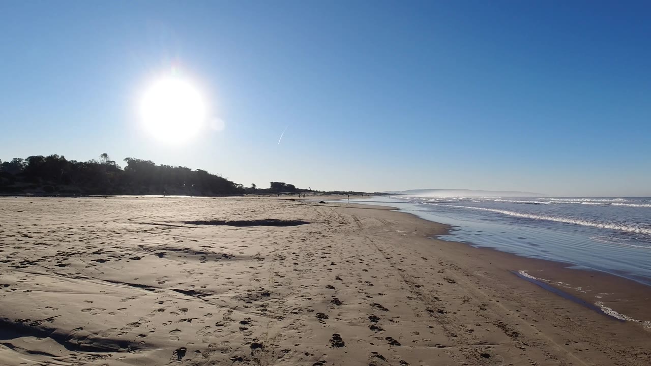 Wide Pismo Beach view with sun glare above the left side, a broad sandy foreground, darker bluff area, and active surf along the right edge, with no visible people.