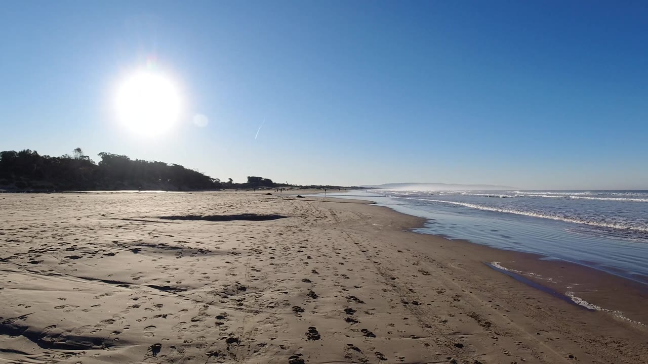 Pismo Beach sand and wet shoreline stretch toward the distance, with bright sun over the left headland and white surf moving along the right, and no visible people.