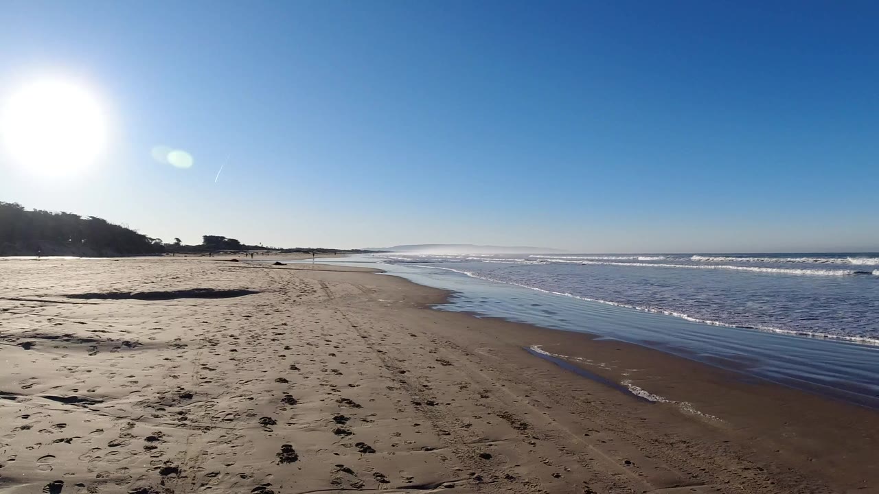 Pismo Beach shoreline at the start of the clip, with dry sand in the foreground, small waves to the right, strong sun flare at upper left, and no visible people.