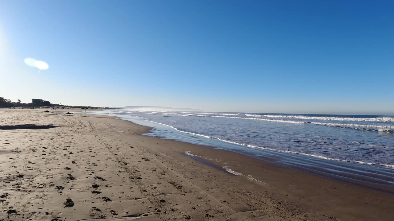 Pismo Beach shoreline at three seconds, with empty sand stretching left, shallow surf along the right edge, bright sky overhead, and no visible people.