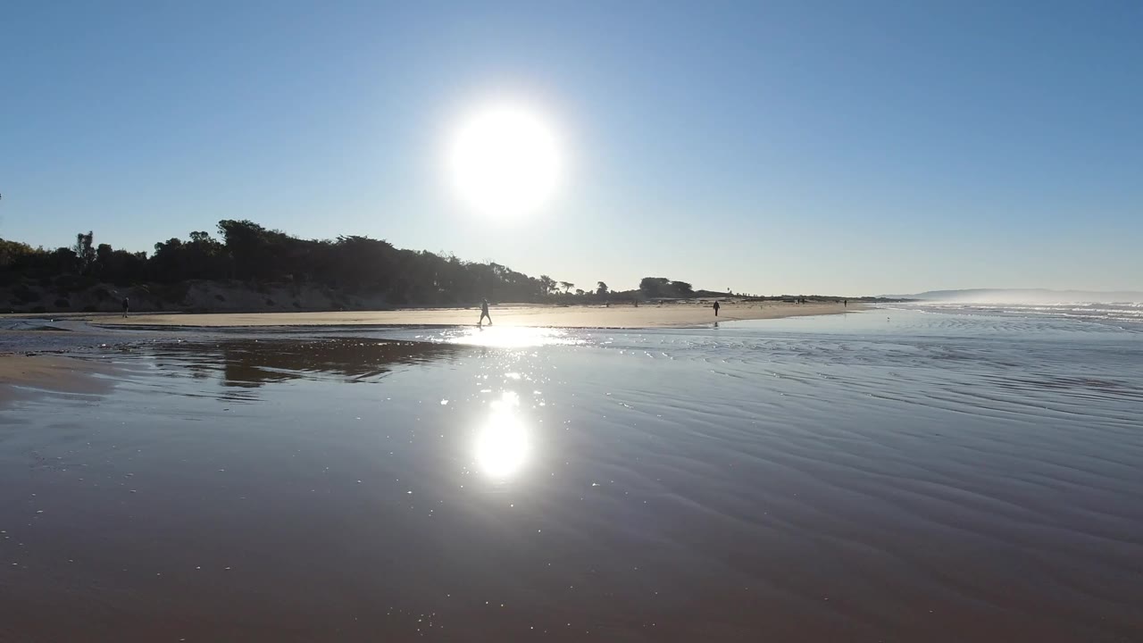 Pismo Beach view one second in, with the sun high over the beach, its reflection stretching across shallow water, and low waves continuing along the right side of frame.