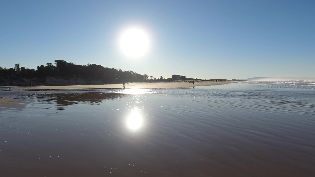 Pismo Beach shoreline at two seconds, with a wide band of wet sand in front, a bright vertical sun reflection, waves edging in from the right, and small figures far along the beach.