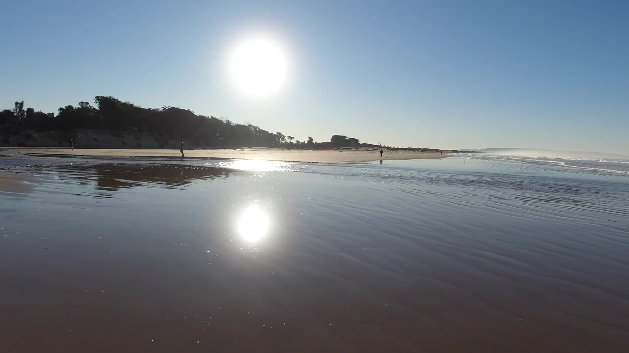 Wide beach view with bright sun, wet sand reflection, surf, and small people walking.