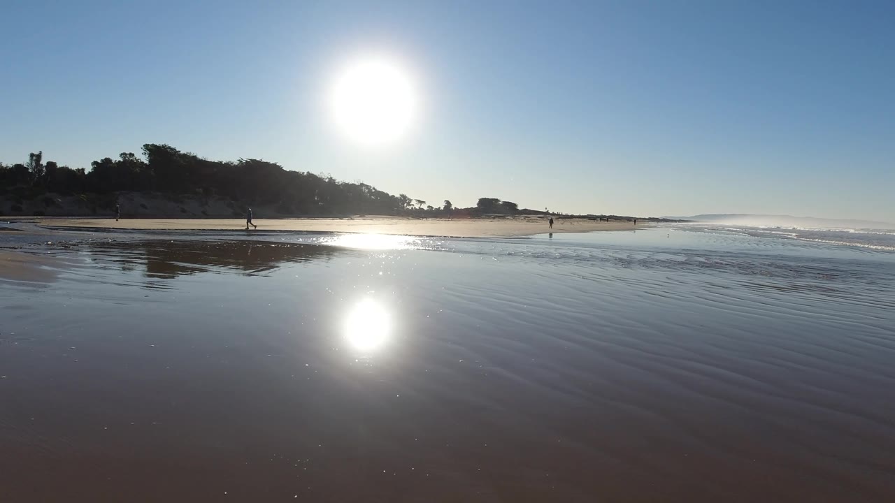 At 0:02, the Pismo Beach horizon stays broad as tiny figures walk near the surf, with bright reflected sunlight on the foreground sand and the darker dune edge on the left.