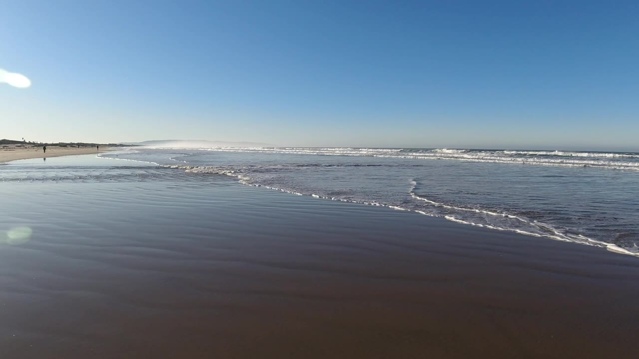 Small waves washing over wet beach sand under a clear blue sky.