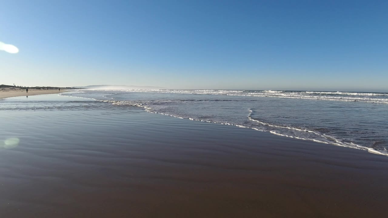 Pismo Beach surf at 0:01 with a curved white foam edge moving across dark wet sand, distant breakers beyond it, and no visible people in the foreground.