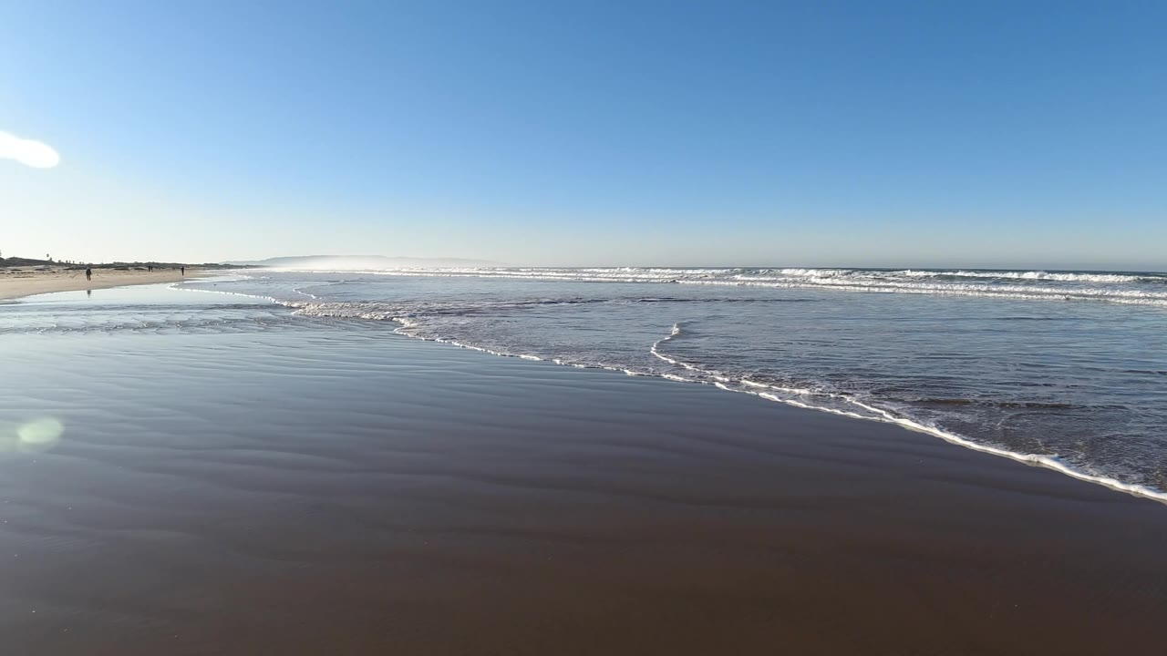 Pismo Beach shoreline at 0:02 with the foam line slightly farther across the foreground, bright glare on the wet beach, distant surf, and no visible people in the foreground.