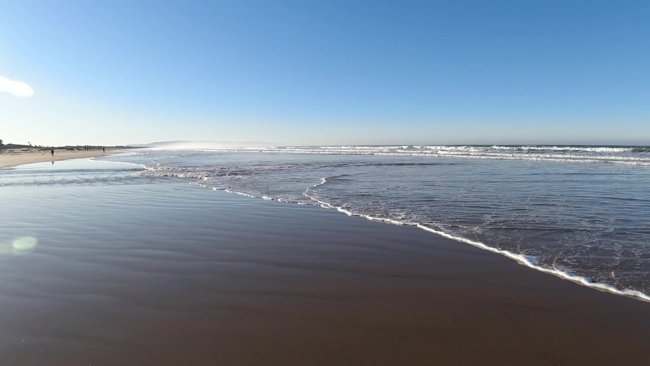 Pismo Beach at 0:03 with a wider sheet of shallow water in the midground, beach stretching from the left edge, distant surf across the horizon, and no visible people in the foreground.