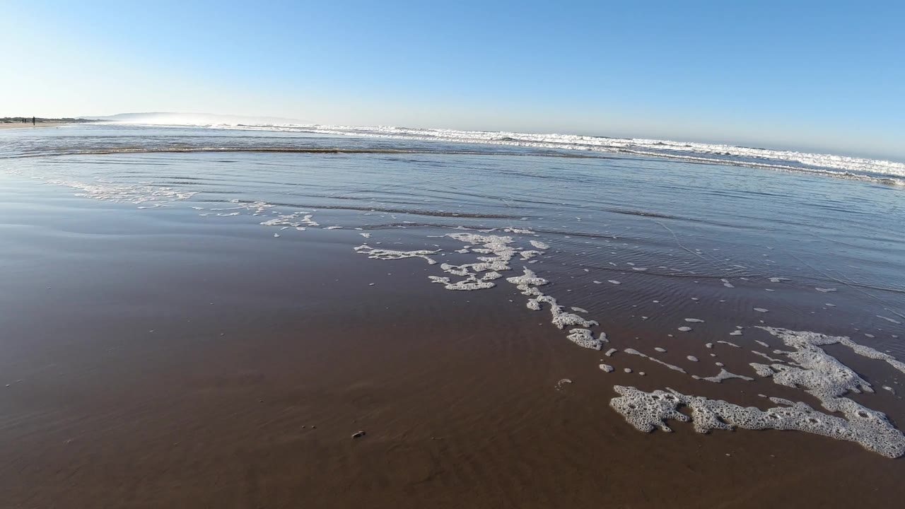 Foam patches sit across reflective wet sand while a broad line of surf breaks in the distance at Pismo Beach, California, with no visible people.