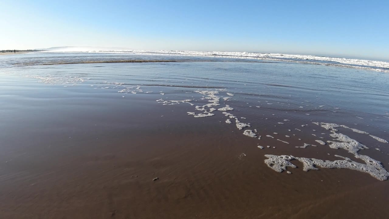 A curved streak of white foam crosses the wet foreground sand toward shallow water and distant waves at Pismo Beach, California, with no visible people.