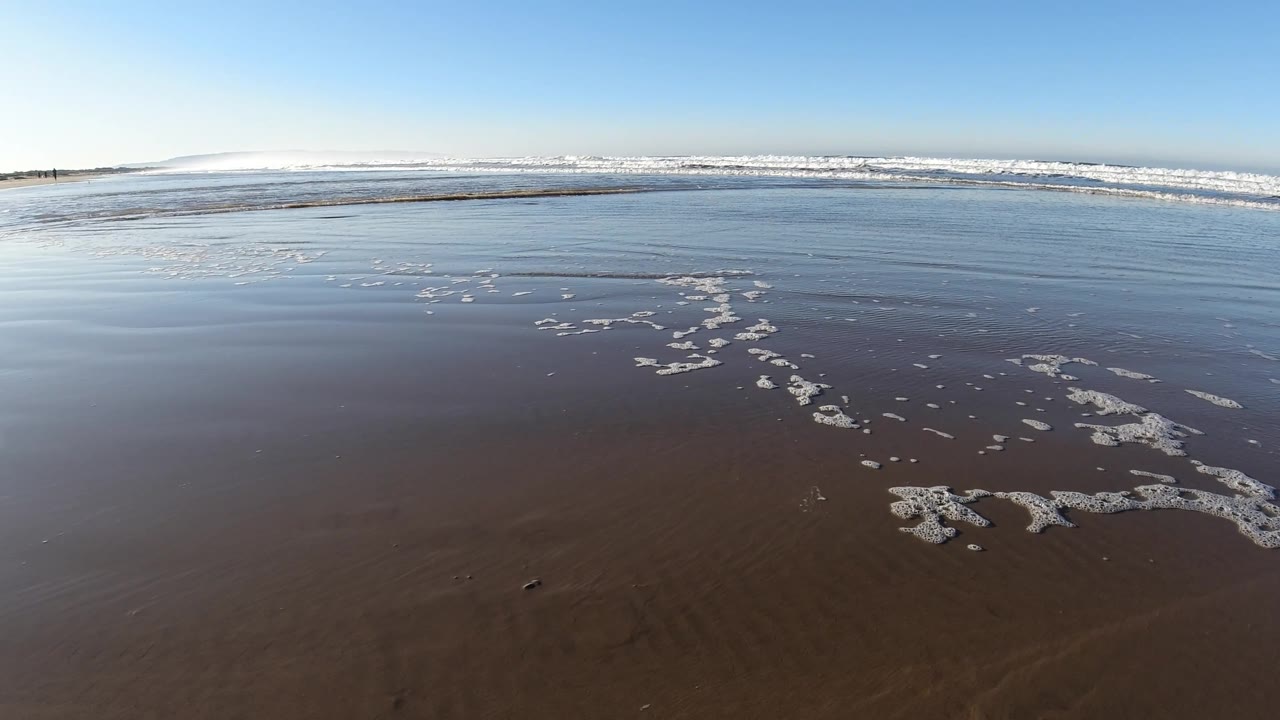 Small foam clusters break up the flat wet shoreline as white surf runs across the background at Pismo Beach, California, with no visible people.