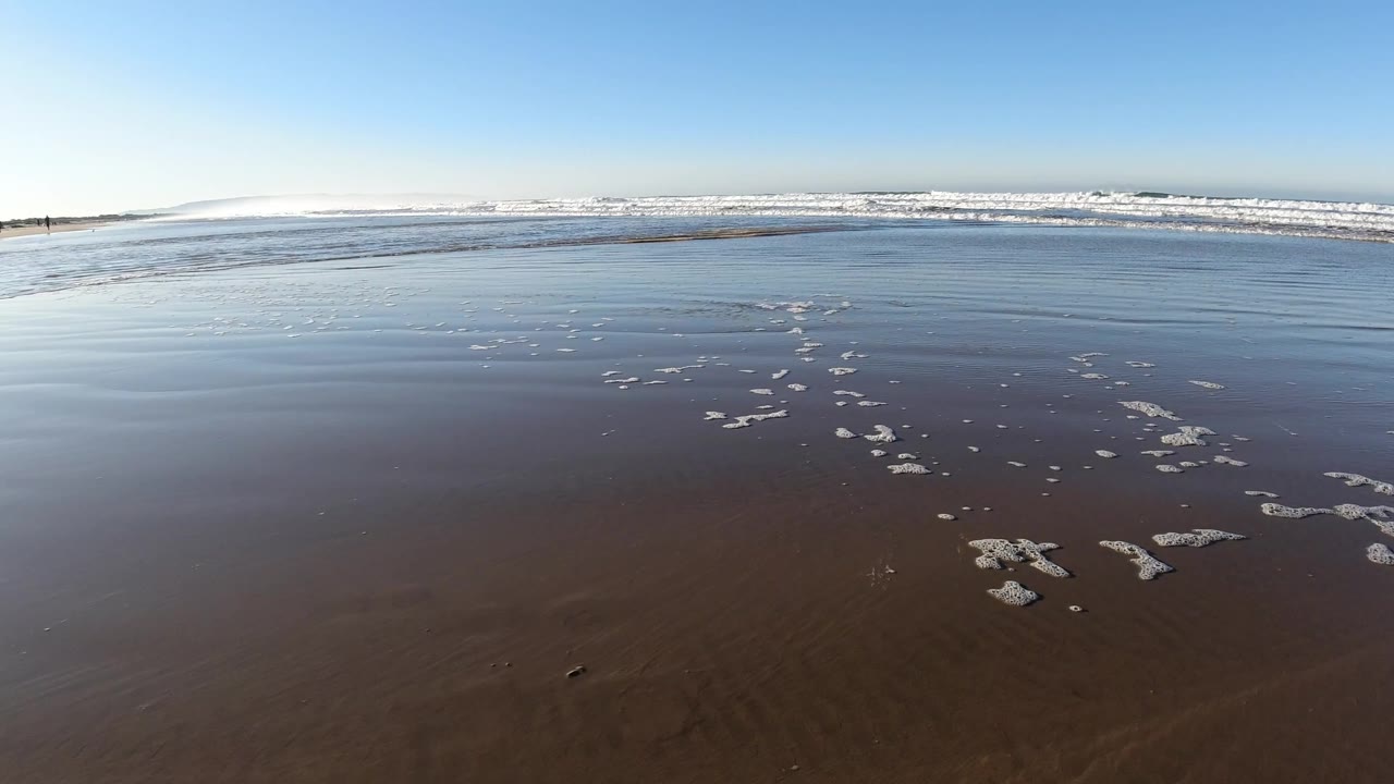 At Pismo Beach, California, small foam clusters sit on smooth wet sand while a broad line of white waves rolls in under a clear blue sky, with no visible people.