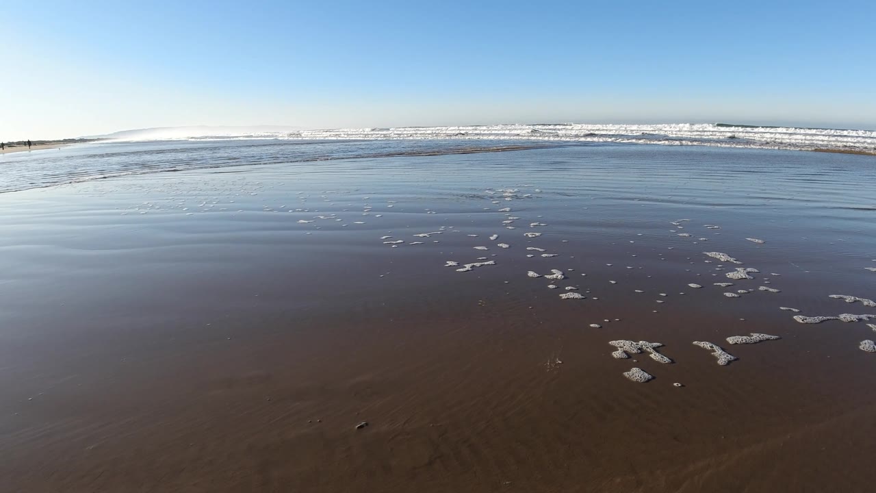 At Pismo Beach, California, the reflective shoreline shows more open wet sand in front, scattered foam to the right, and breaking waves across the background, with no visible people.
