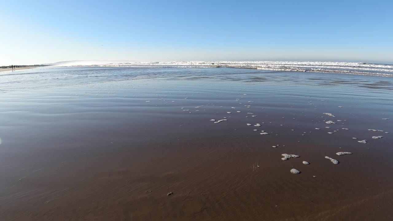 A thin sheet of water glides over the Pismo Beach sand while small foam patches sit near the lower right and low waves roll across the background, with no visible people.
