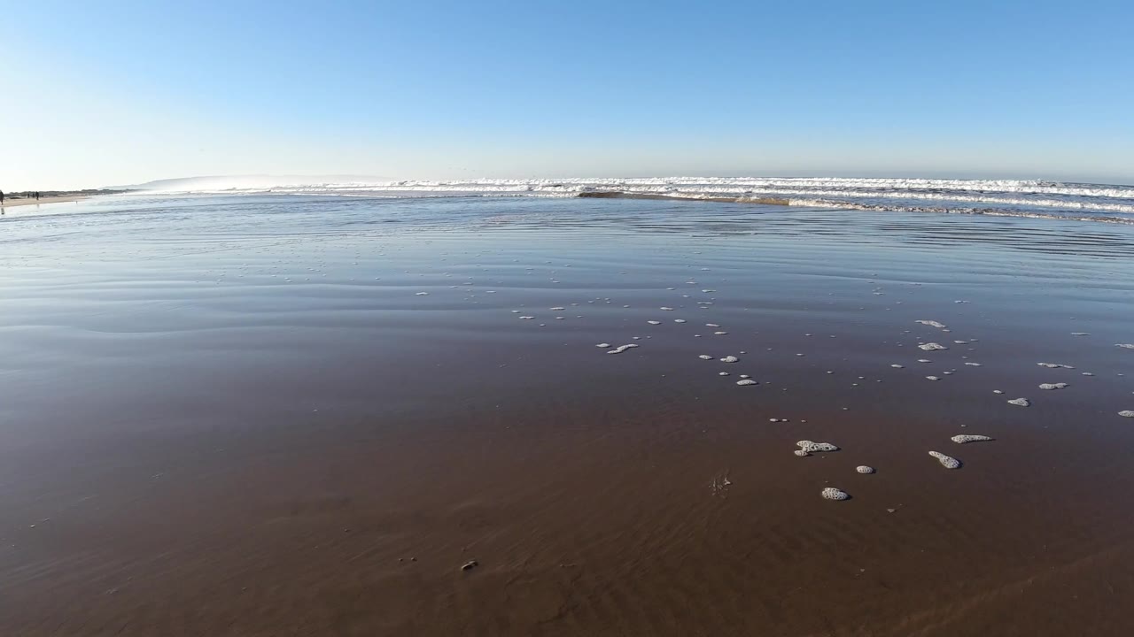 Pismo Beach wet sand reflects shallow water as small foam patches sit in the foreground, surf breaks near the horizon, and no visible people appear.