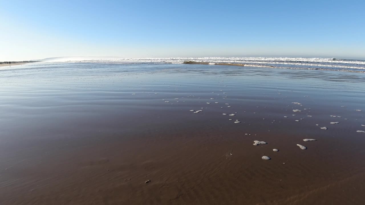 At 0:02, small white foam clusters dot the lower frame while smooth wet sand leads to a band of breaking surf at Pismo Beach, with no visible people.
