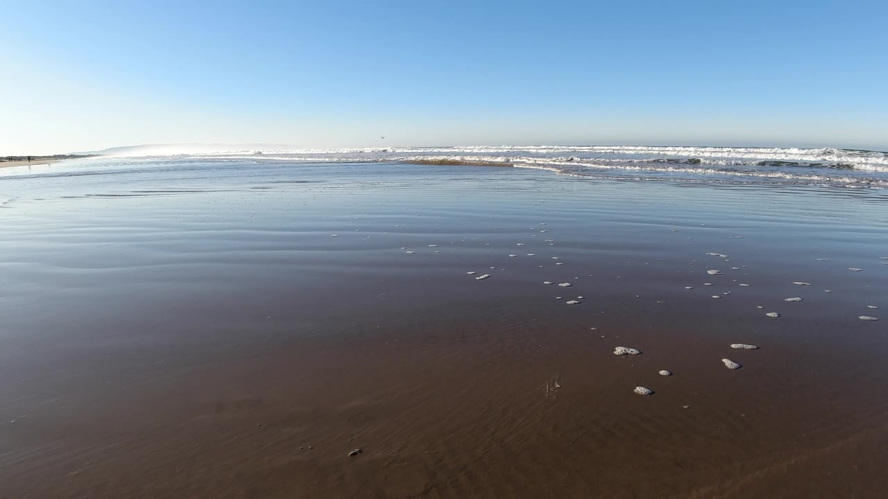 Wide Pismo Beach low-tide view with a broad band of smooth wet sand, shallow blue-gray water, white surf in the distance, and no visible people.