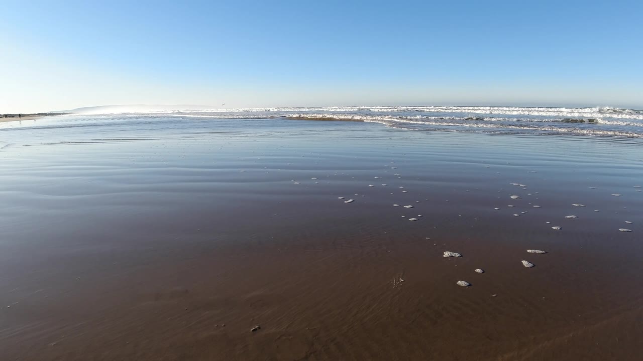 Pismo Beach beach flats with wet brown sand in the lower frame, pale reflective water in the middle, a thin surf band near the horizon, and no visible people.