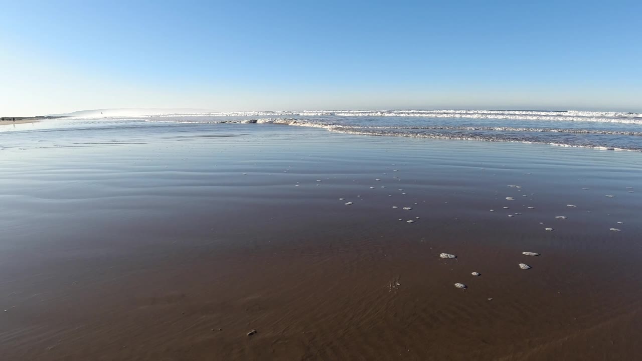 Pismo Beach shoreline at 0:00 with reflective wet sand, scattered foam spots, low surf across the background, and no visible people.