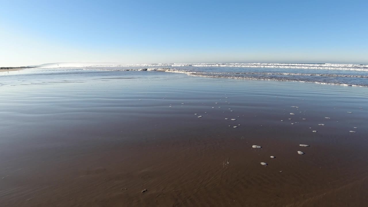Pismo Beach at 0:01 shows shallow reflective water over brown sand, a line of white surf near the horizon, and no visible people.