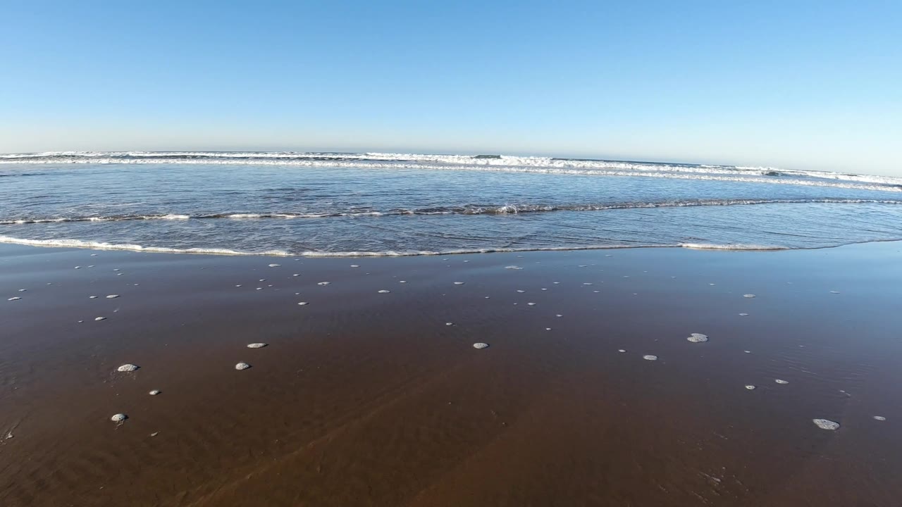 A slightly shifted Pismo Beach shoreline view shows more reflective water over the sand, thin surf bands, a bright blue sky, and no visible people.