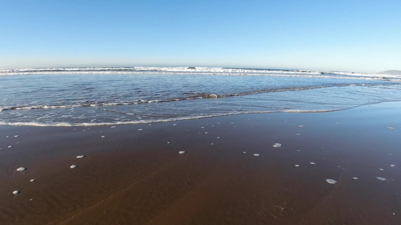 Pismo Beach surf reaches across the lower left, with dark wet sand in front, low rolling waves beyond, distant land faintly at the right horizon, and no visible people.