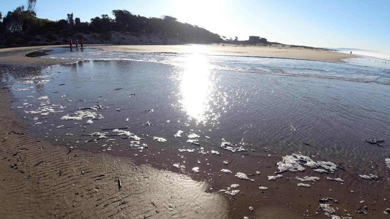 The sun reflection runs down the center of the shallow shoreline at Pismo Beach, California, with scattered foam patches in front and tiny people standing near the left sandbar.