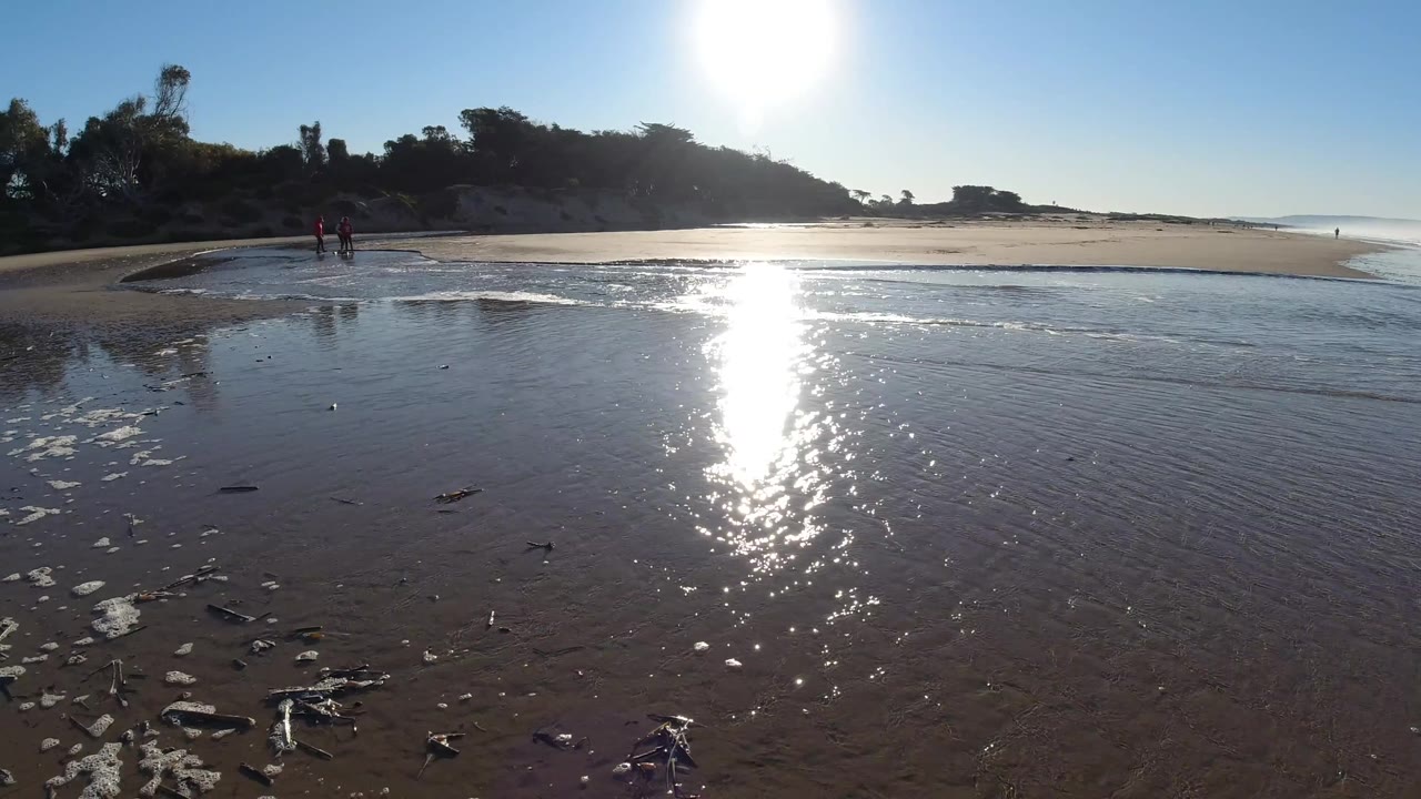 A wide Pismo Beach shoreline view shows rippled wet sand and scattered debris in the foreground, a vertical strip of sun glare on tidal water, and two distant walkers left of center.