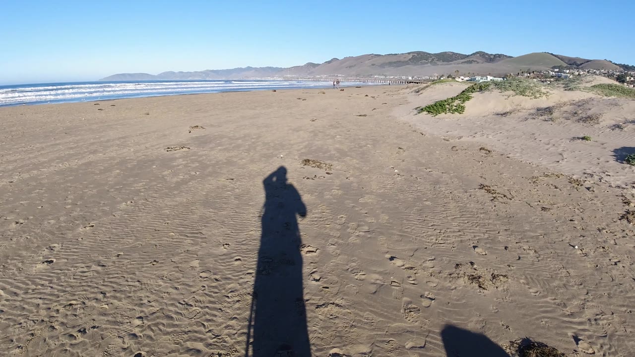 Wide wet sandy beach with waves at left, dunes at right, and a long human shadow in the foreground.