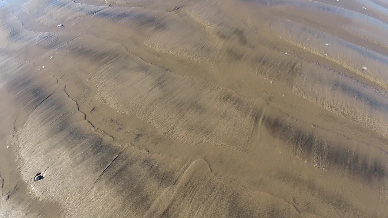 Second overhead frame at Pismo Beach showing wavy wet sand ridges, darker mineral-like streaks, small shells, and no visible people.
