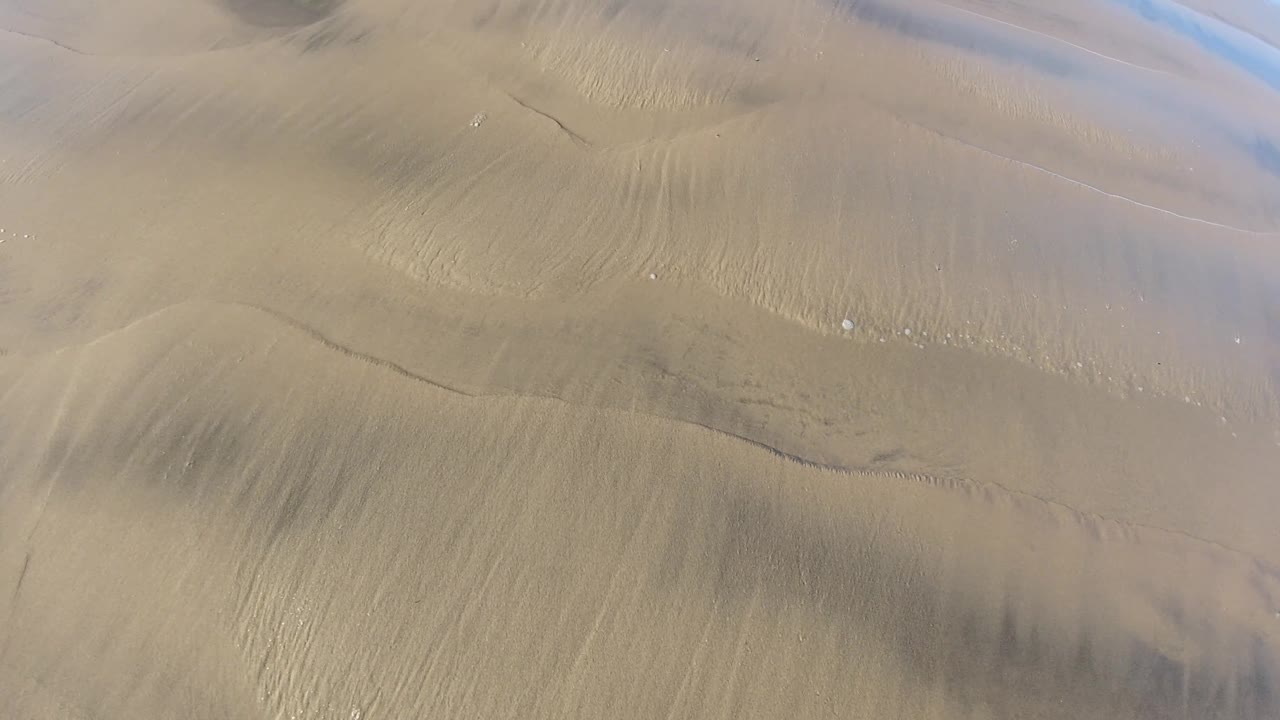 Overhead Pismo Beach, California view of dry rippled ground where dark cracks and a broad shadowed band run through the sandy surface, with no visible people.