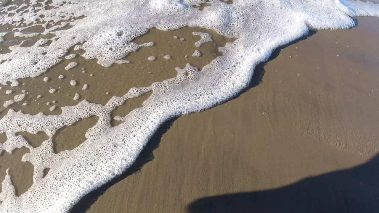 Foamy ocean water reaches across wet sand at Pismo Beach, California, with a dark human shadow falling into the lower part of the frame and no visible face or body.