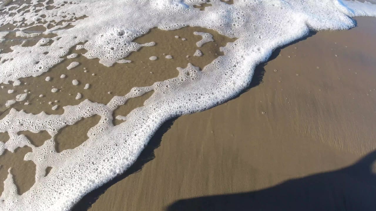Foam line curving across wet beach sand with shadow at the bottom edge